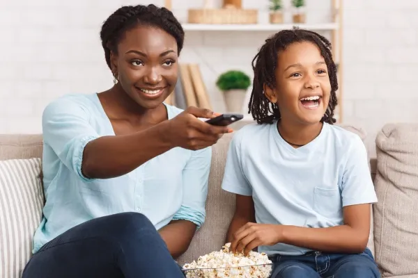 A smiling mother and her son are sitting on a tan sofa, with the mother holding a remote control and the son happily laughing while holding a bowl of popcorn.